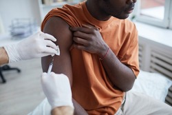 Close up of adult African-American man looking away while getting covid vaccine in clinic or hospital, with male nurse injecting vaccine into shoulder
