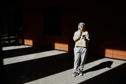 Graphic wide angle portrait of modern Middle-Eastern woman using smartphone in city lit by sunlight and wearing headphones, copy space