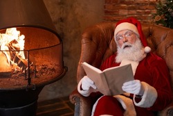 Curious senior Santa Claus in eyeglasses sitting in comfortable armchair at fireplace and resting with book