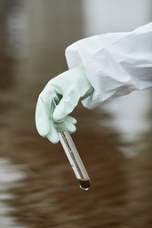 Close up of unrecognizable scientist wearing hazmat suit collecting water samples, focus on gloved hand holding test tube