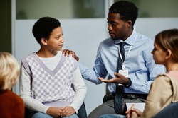 Portrait of young African-American psychologist supporting teenage girl in group therapy meeting