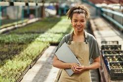 Pretty young African American female farmer or woner oh large hothouse with laptop standing against long tables with seedlings