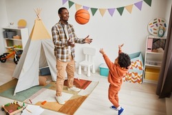 Full length portrait of young black father playing with toddler son indoors and throwing basketball ball in cozy kids room interior