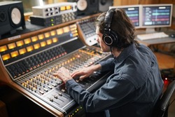 Portrait of young man wearing headphones and operating buttons and toggles at digital audio workstation in recording studio, music production concept