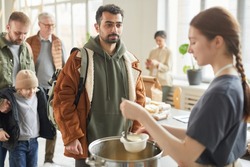 Portrait of young man standing in line at soup kitchen with young woman giving out simple meals to people in need