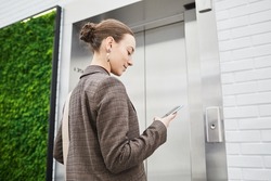 Minimal low angle shot of young businesswoman waiting for elevator in modern office building and using phone, copy space