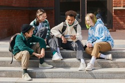 Group of teenage students sitting outside school buildings and watching funny video on smartphone