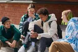 Group of friends sitting on steps of building outdoors and laughing while watching funny video on mobile phone