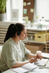 Young woman in headphones sitting at her workplace with laptop and talking to customers online