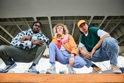 Group of three young people wearing street style clothes outdoors while sitting on stairs in urban area and looking at camera
