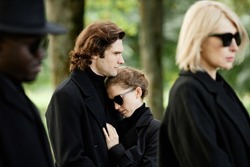 Portrait of young couple wearing black grieving together and embracing at outdoor funeral ceremony