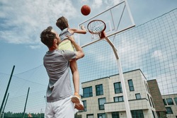 Low angle portrait of father and son playing basketball together, man holding boy shooting ball through hoop