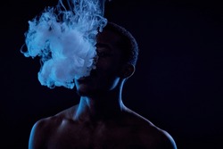 Young African American man standing in front of camera in darkness and blowing cloud of vapor out of his mouth while smoking