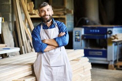Portrait of mature bearded carpenter posing confidently with arms crossed standing in woodworking shop and smiling happily looking at camera