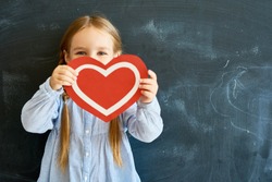Portrait of  adorable little girl hiding her face behind  red paper heart and looking at camera  posing against blackboard in school