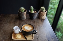 A hot cup of cappuccino served with cookies and sugar on wooden tray setting on wooden table next to window with green plant view and cactus and black wall background 