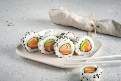A set of fresh sushi rolls with salmon, avocado and black sesame seeds served on a plate with chopsticks. Japanese sushi uramaki or California roll