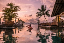 Luxury swimming pool in a tropical resort, relaxing holidays in Seychelles islands. La Digue, Young man during sunset by swim pool, men watching sunset in infinity pool
