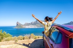woman outside car window road trip with hands up, car at Chapmans Peak Drive in Cape Town South Africa looking out over the ocean. 