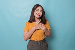 Happy mindful thankful young woman holding hands on chest smiling isolated on blue background feeling no stress, gratitude, mental health balance, peace of mind concept.