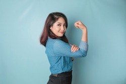 Excited Asian woman wearing a blue shirt showing strong gesture by lifting her arms and muscles smiling proudly