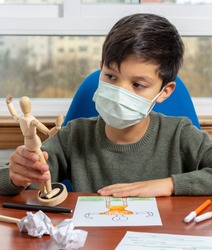 Child with mask observing a wooden human figure to copy and draw it. Some crumpled sheets of paper on his desk
