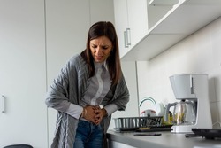 Unhappy sick woman with hands holding pressing her crotch lower abdomen. Medical or gynecological problems, healthcare concept. Mid adult woman suffering from abdominal pain while standing in kitchen.