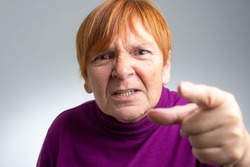 Hey you! Senior woman pointing at camera finger with serious face. Emotion and feelings concept. Portrait of expressive grandmother standing with collected red hair. Isolated on gray background.
