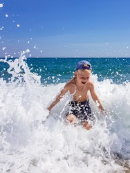 A little blond boy is playing on the seashore, laughing, running away from the waves against the background of huge waves and splashes, Calabria, Italy