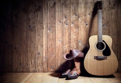 Acoustic guitar, cowboy hat and boots resting against a blank wooden plank grunge background with copy space