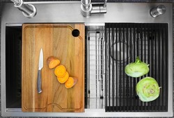 Workstation kitchen sink with cutting board and draining rack used to slice a sweet potato and to dry kohlrabi vegetable