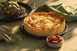 Golden organic healthy crispy pie quiche with artichokes, dried tomatoes and olives on the table, evening sunlight in the summer garden, soft selective focus
