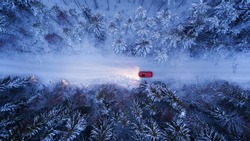 A red car rides along a winter road in the night forest. Snow on trees and roadsides, Aerial View.