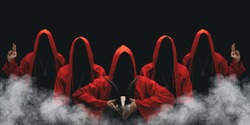 Group of mysterious figures in hooded cloaks in the dark. Leader of sectarians holds skull with horns. Horror scene with smoke. Black background.