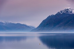 Tranquil sunset over Lake Hawea, New Zealand. Mist hangs over the mountains with still lake in the foreground