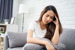 Beautiful Asian woman wearing white T-shirt feeling stress and headache on sofa. She used her hand to touch her face. Office syndrome concept.
