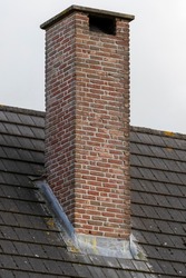 An isolated portrait of a tall masonry grout red brick chimney on a black tile roof on an overvast day.
