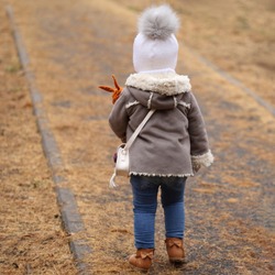Backview of little girl in lightbrown short sheepskin coat, holding doll in park on cold day.