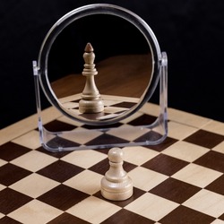 An ambitious chess pawn on a chessboard looks in the mirror and sees himself as a queen or king. The concept of confidence, ambition, self-belief,success,business.