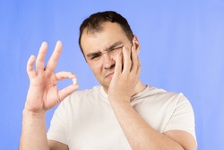 Man in a white T-shirt on a blue background holds a wisdom tooth in his hands after surgical tooth extraction.Man after an operation to remove wisdom teeth.Pain in wisdom teeth, concept of dentistry