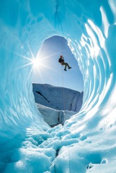 Active young man ice climber on glacier rappelling into ice cave entrance in Alaska.