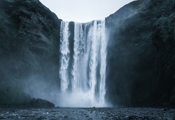 Famous Skogafoss Waterfall in Iceland from the front with rough looking water falling off the hill in a dark look with black stones in the foreground and no people in front during night