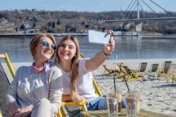 Mother and daughter spending quality time together, bonding, drinking coffee, and taking a selfie by the riverbank with the bridge in the background.