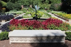 Photograph of large concrete decorative stone bench in front of colorful landscaped garden of plants and flowers during the day in the sun light. Full color photo.