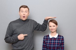 Studio shot of surprised tall man showing height of short woman, pointing at her with finger, both are standing over gray background. Concept of diversity of people's heights, tall and short persons
