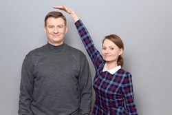 Studio shot of short woman standing on tiptoes and showing height of tall man, both are cheerful and smiling, over gray background. Concept of diversity of people's heights, tall and short persons