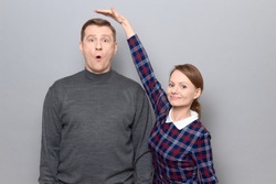 Studio shot of short woman standing and showing height of tall man, woman is smiling, man is amazed, over gray background. Concept of diversity of people's heights, tall and short persons