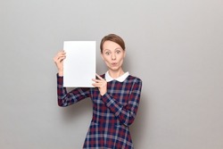 Studio portrait of surprised amazed young blond woman holding white blank paper sheet with place for your text and design in hands, wearing checkered dress, standing over gray background