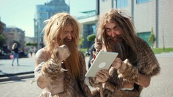 Excited prehistoric bushmen of hunter-gatherers covered in fur browsing internet on tablet discovering technology in modern city. Adaptation concept.