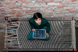 Elevated view of elementary age cheerful School girl of Indian Ethnicity sitting  on cot holding chalkboard wearing school uniform. She is looking up to the camera while sitting on the bed
